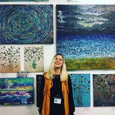 Krissie standing in front of a gallery wall mounted with her paintings